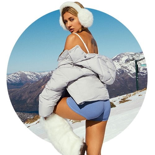 Model poses on a ski hill wearing white fur earmuffs, a grey puffer off around her shoulders, blue shorts, and white boots.