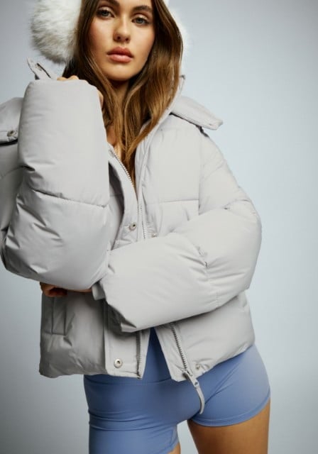 Model wears white earmuffs, a grey puffer, jeans, and white boots.