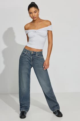 Model wears a white off the shoulder top and mid-wash baggy jeans with black shoes.