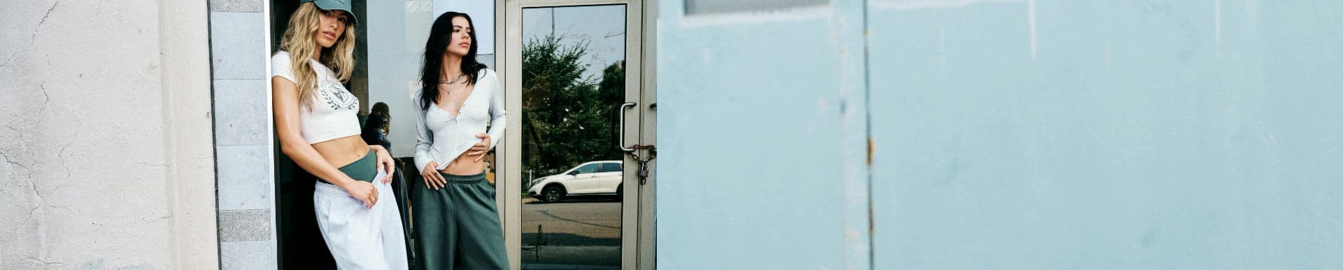 Two models poses by a door. One wears a green baseball cap, white graphic tee, and baggy grey sweats with shorts underneath. The other wears a low cut white long sleeve shirt and baggy green sweats. 