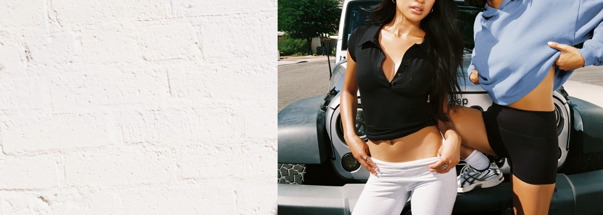 Two models pose by a car. One is wearing a black polo with the buttons undone and white pants. The other is wearing a blue hoodie and black shorts. 