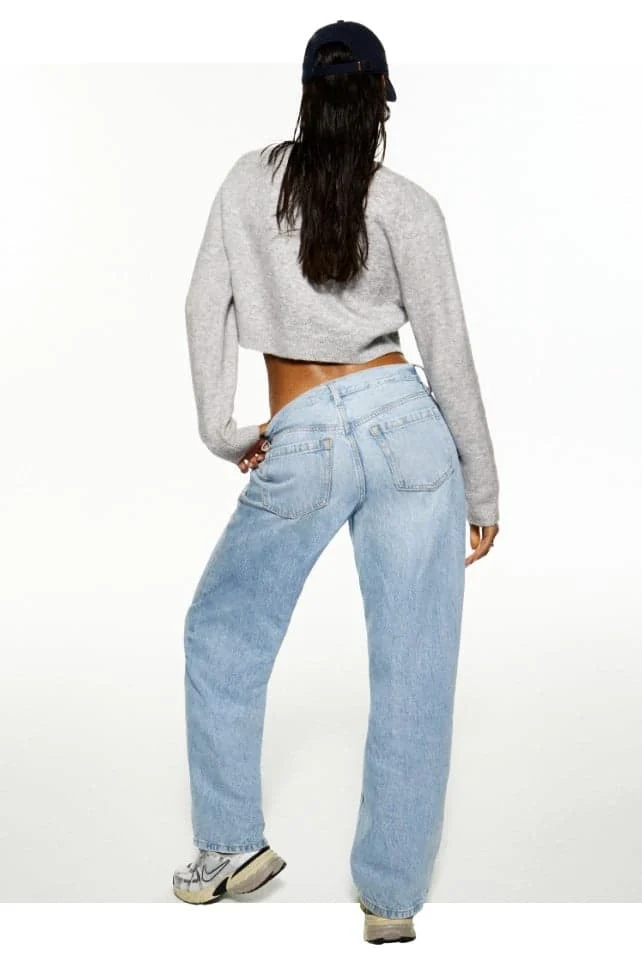 Model poses in a grey cardigan with no shirt and baggy light wash jeans. 