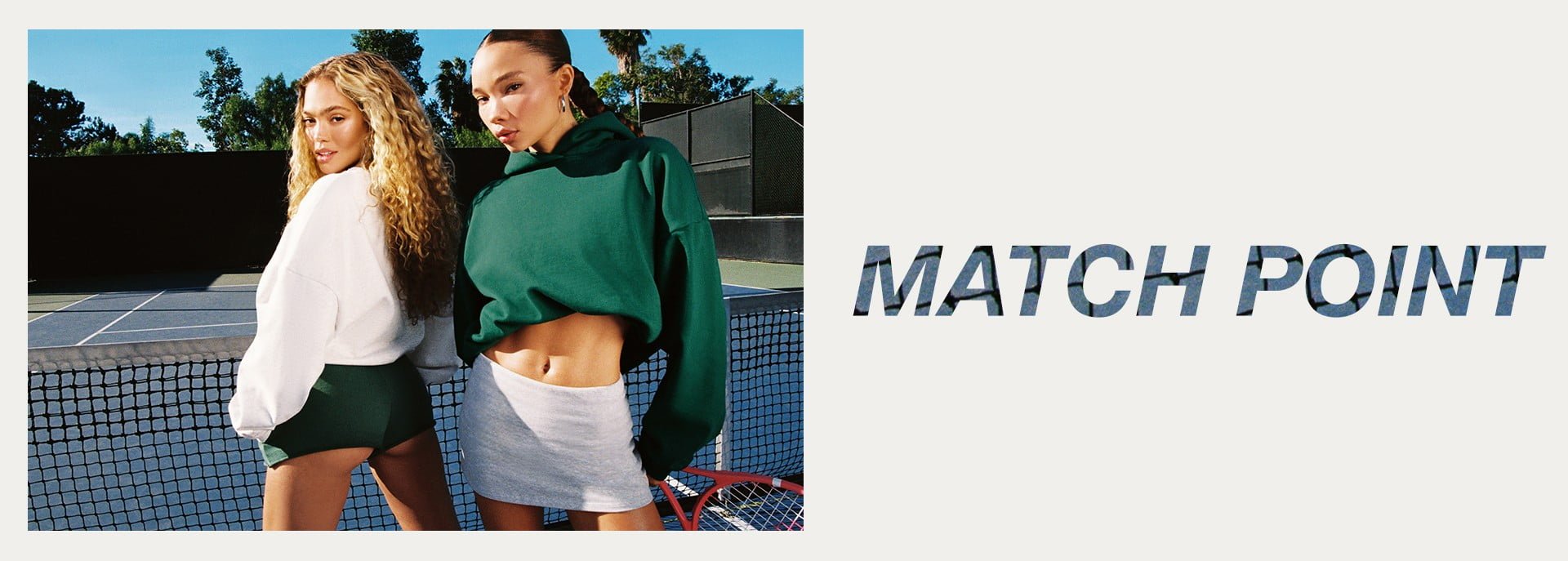 Two models pose on a tennis court, one in a white sweatshirt and green shorts, the other in a green hoodie and white skirt.