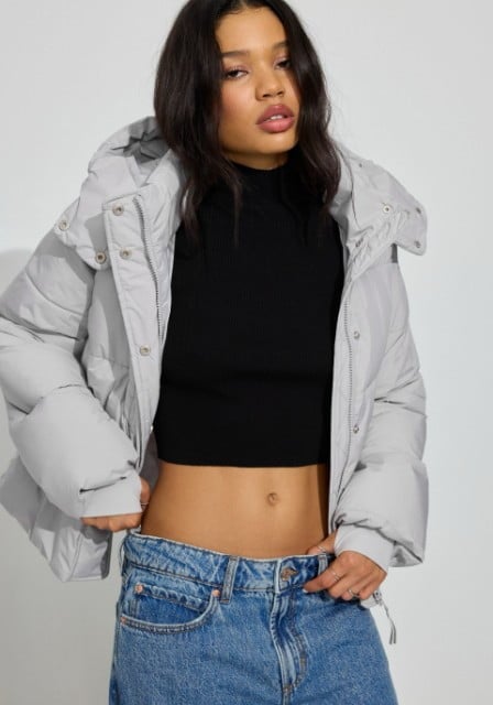 Model poses in a cropped black  top, light grey puffer coat, and jeans.