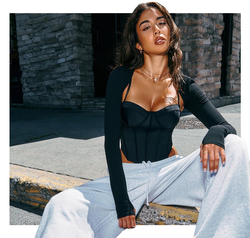 Model sits on the sidewalk wearing a black corset top, a black shrug, and light grey sweats.